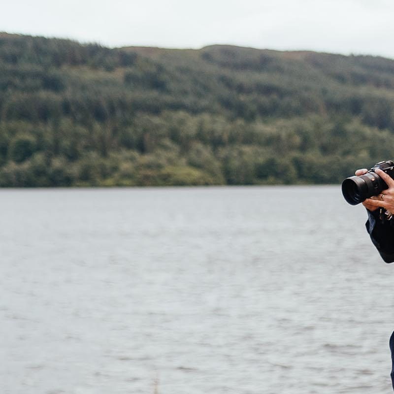 A man stands with a camera filming by Venachar Lochside