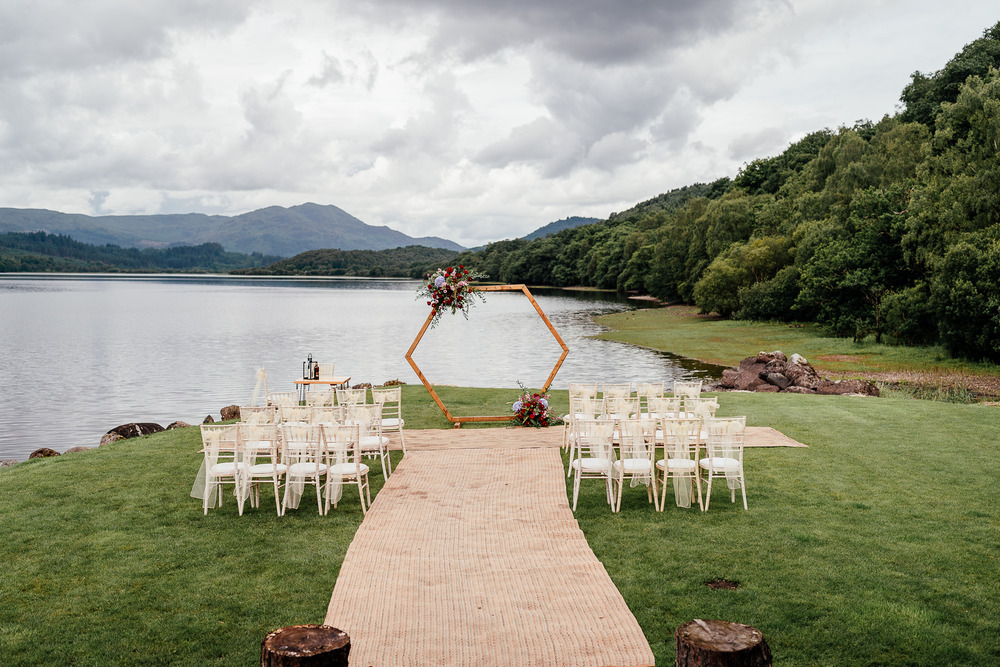 Beautiful view of a wedding ceremony on the lochside. Hexagon arch at the end of the aisle with cream seats for guests. Hessian aisle to walk down to the loch edge. Mountains in the background are overlooking the loch.